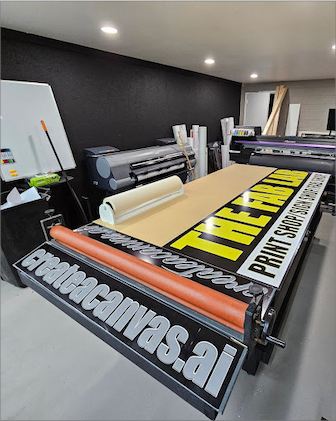 A well-organized print shop with large format printers and rolls of paper in the background. In the foreground, a custom sign with bold yellow lettering on black background reads 'THE FAB LAB PRINT SHOP' on a flatbed cutter. Below, a banner features 'printedcanvas.ai' in white lettering on a black background.